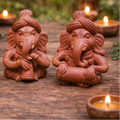 Three terracotta Ganesh statues on a wooden surface with lit candles in the background.
